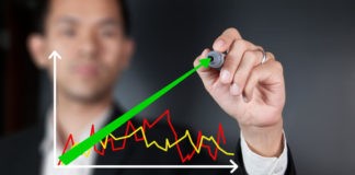 Wibest Broker- Stock Exchanges: A hand holding a pen while shares are being reviewed showing an increase in the stock market.