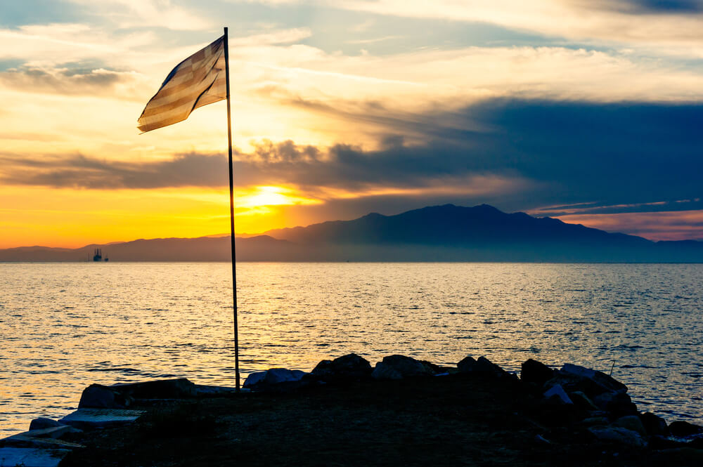 Greek flag waving on shoreline at sunset.