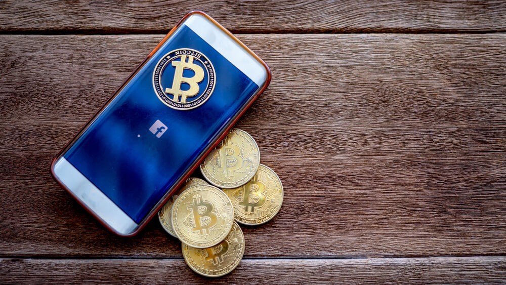 Wibest – Digital Coins: Mobile phone with symbol of Facebook and digital money Bitcoin coin on the wooden floor.