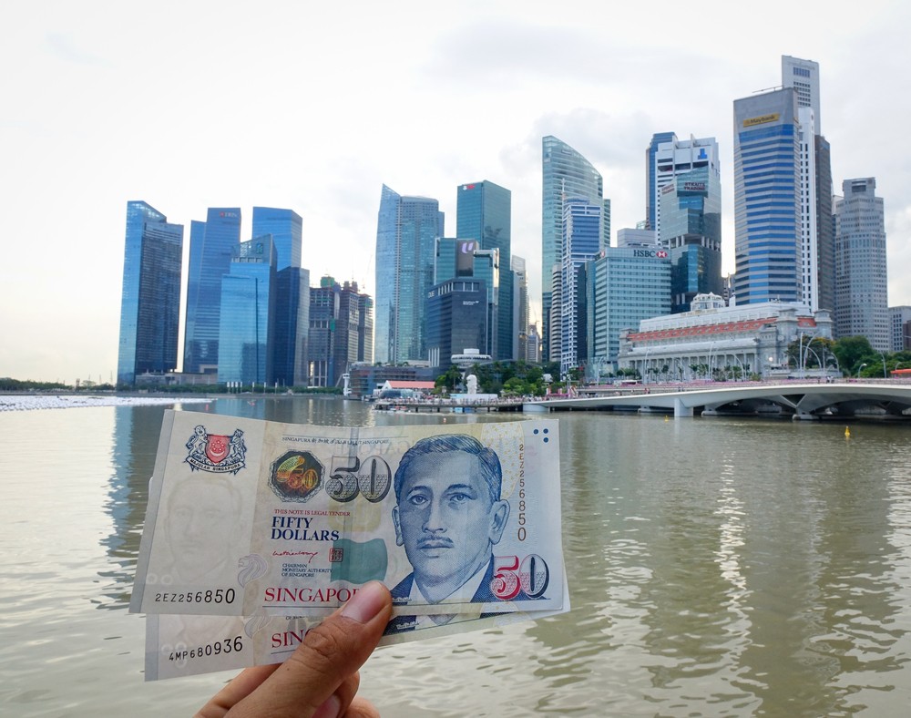 Wibest – Monetary Authority of Singapore: Singaporean dollar in held by a man and a city in the background.