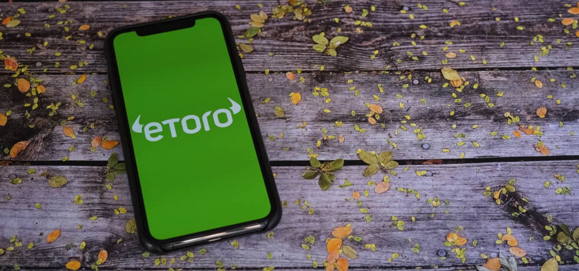 Etoro on a smartphone screen on an old wooden table with leaves on it.