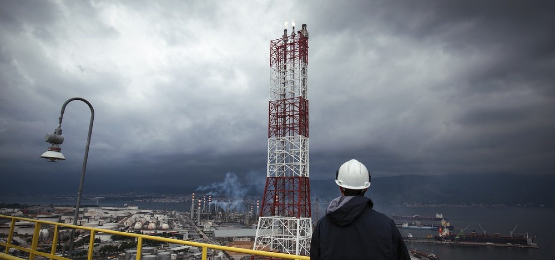 Wibest – Climate Week: A man staring at an oil refinery under a stormy sky.